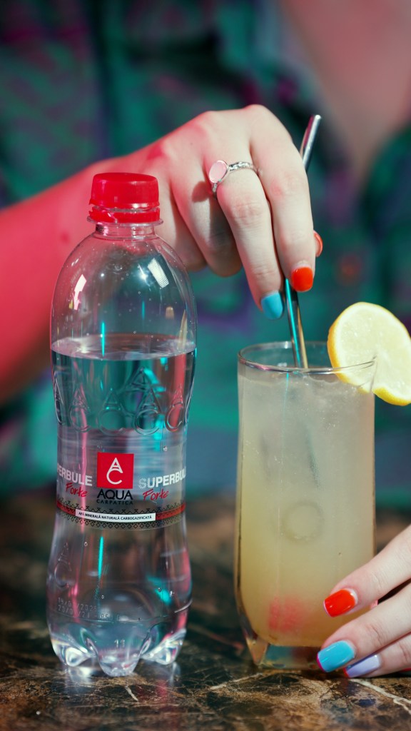 mână de femeie ține un pai intr-un pahar inalt cocktail tom collins cu lamaie si apa minerala aqua carpatica forte