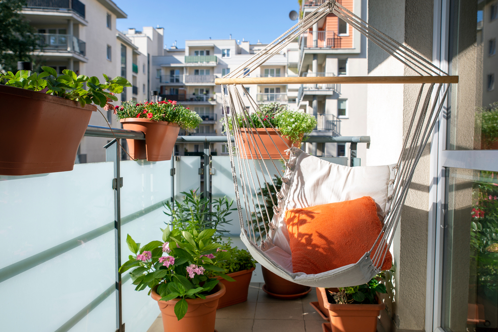 Un balcon urban însorit cu flori de primăvară în ghiveci și un fotoliu suspendat ca mobilier.