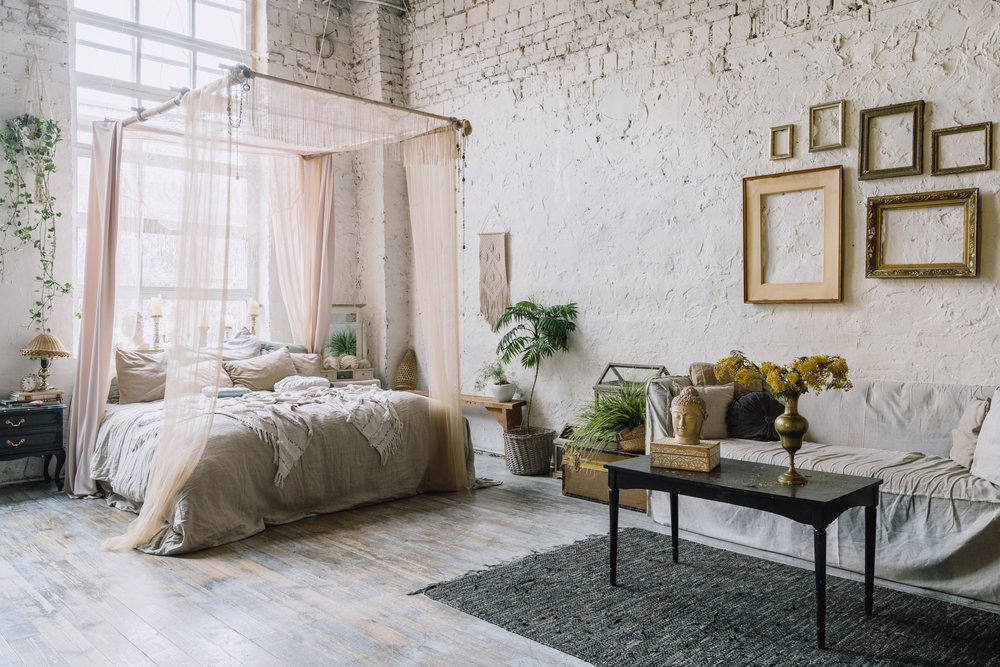 Un dormitor de tip loft, cu pereți din cărămidă vopsiți în crem deschis, cu un pat cu baldachin amenajat în culori calde, măsuță din lemn negru și decorațiuni aurii.