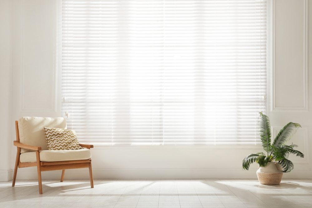 Draperii sau jaluzele, ce să alegi pentru casa ta: o cameră luminoasă cu draperii albe transparente la geamuri, și draperii, un fotoliu retro și un palmier în ghiveci.
