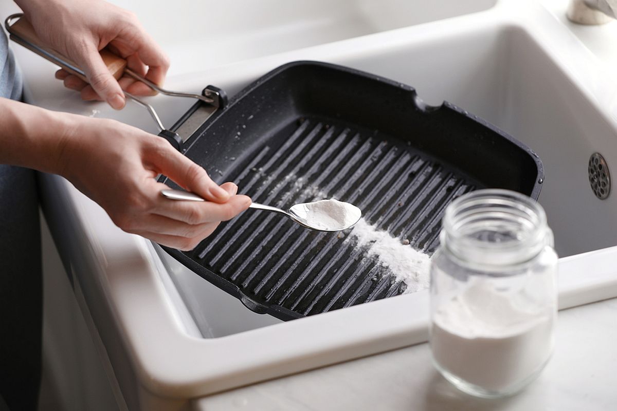 Mîini de femeie presară bicarbonat de sodiu pe o tigaie grill neagră ca s-o curețe.