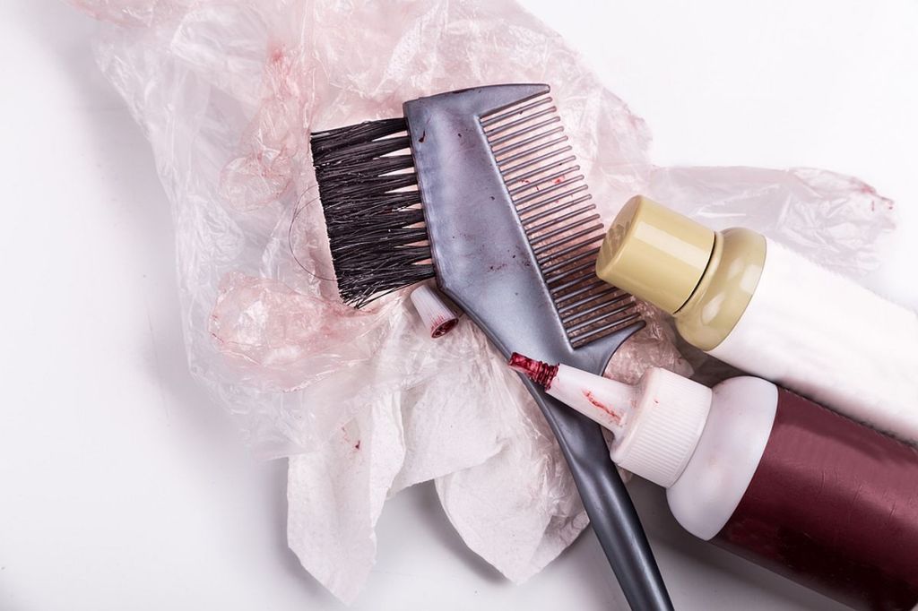 Pete vopsea de păr pe haine: Un tub de vopsea de păr roșie, alături de un recipient alb, mănuși din plastic și perie pentru vopsit.