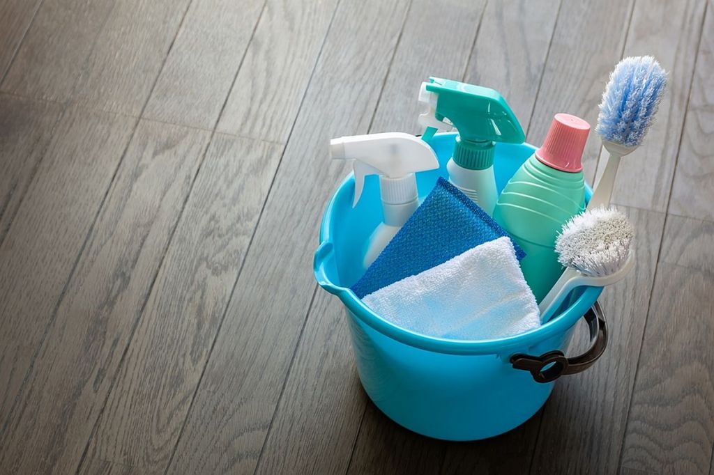 Cu ce se curăță lavabila de pe parchet: o găleată albastră cu recipiente cu pulverizator și lavete pentru curățenie în ea, pe parchet maro.