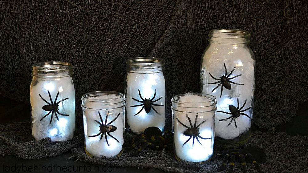 Decorare casa Halloween 2024: Borcane transparente umplute cu pânză albă, luminițe și pâianjeni negri puse pe un fundal negru din material textil.