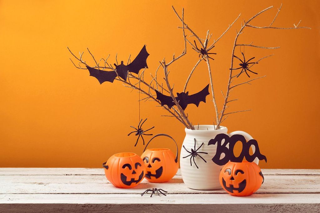 Decorare casă Halloween 2024: Dovleci de Halloween portocalii din plastic, alături de o cană albă cu pâianjen negru, în care sunt puse crenguțe negre cu lilieci, pe o masă din lemn natur și fundal portocaliu.