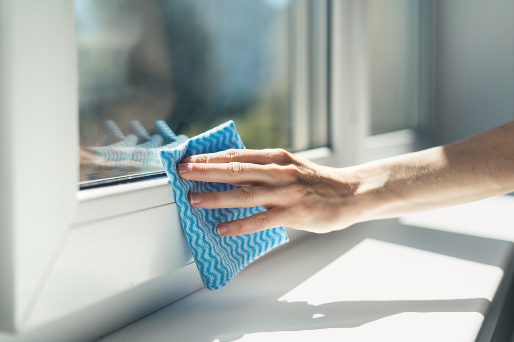 Cu ce se curăță lipiciul de pe termopane: o mână de femeie curăță cu o lavetă geamul termopan, în lumina directă a soarelui.