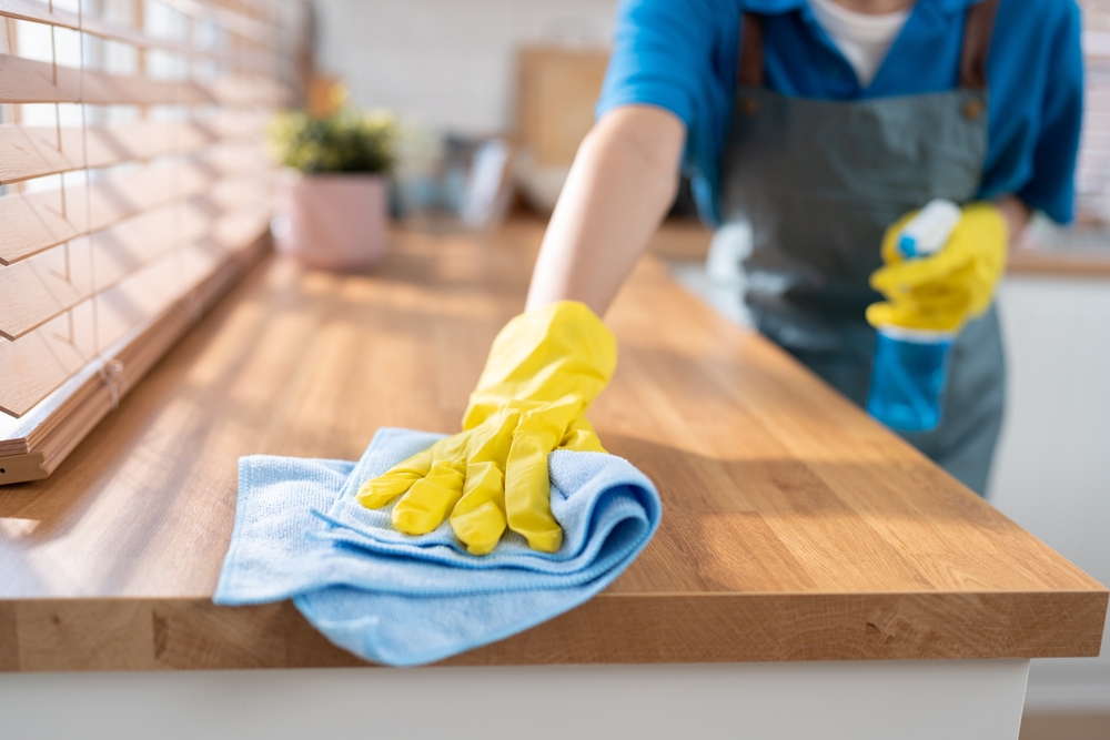 Cu ce se curăță mobila din bucătărie din PAL de grăsime: o mână cu mănușă de menaj galbenă șterge blatul de bucătărie cu o lavetă din microfibră bleu.