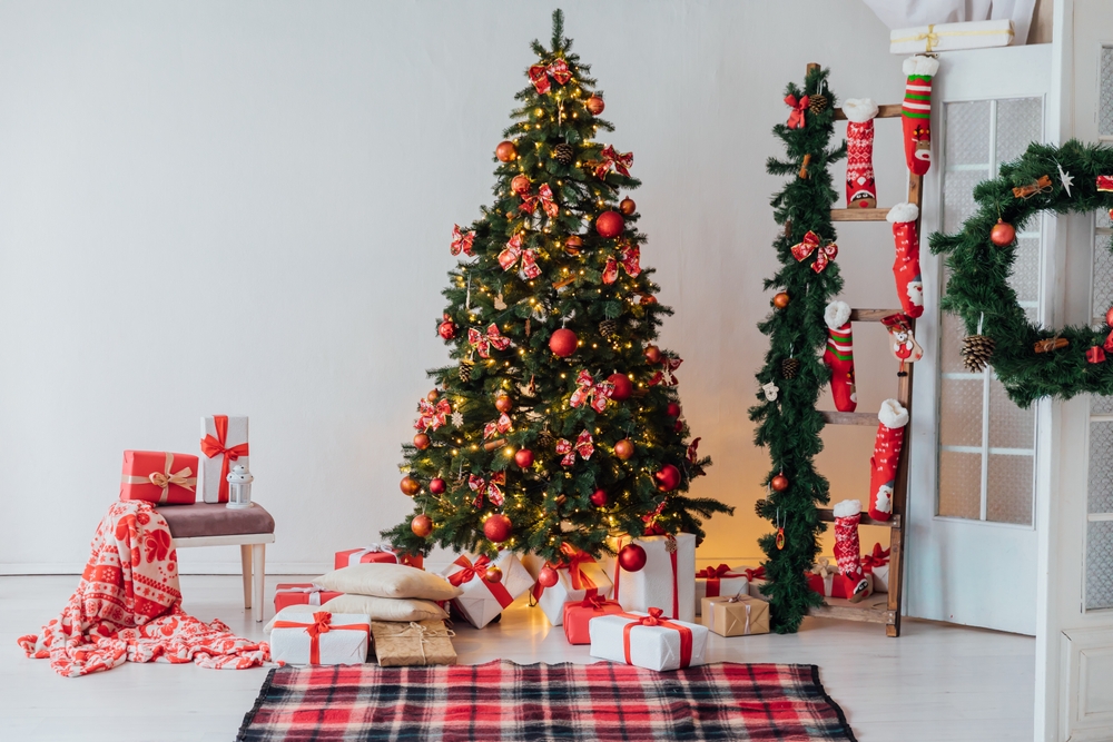 brad de craciun impodobit cu decoratiuni rosii, in living, cu cadouri si alte decoratiuni festive de iarna