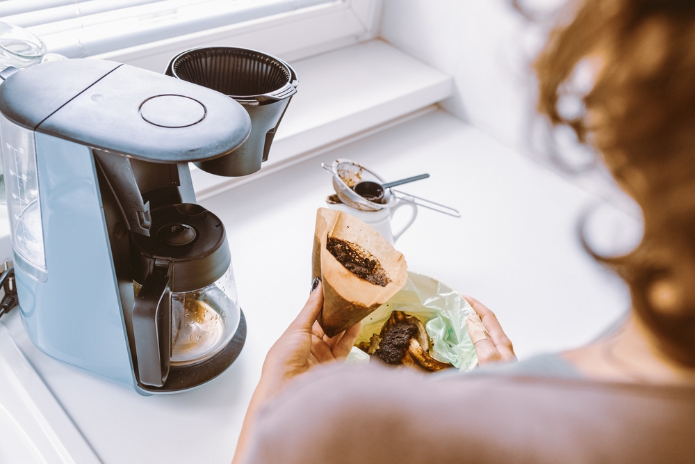 Curățare cafetieră cu oțet: o femeie curăță o cafetieră pe pervazul unei ferestre în interior, aruncând zațul din filtru.