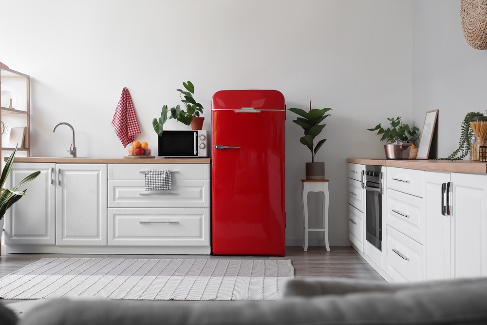 Distanța între frigider și calorifer: Un frigider roșu într-o bucărătie cu corpuri de mobilier din lemn alb și plante.