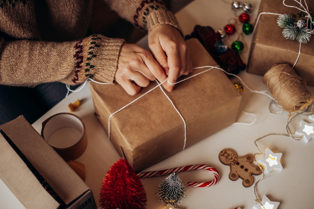 Mâini de femeie îmbrăcată într-un pulover maro deschis cu modele geometrice negre la mâneci leagă cu sfoară albă un cadou ambalat în hârtie de cadouri maro.