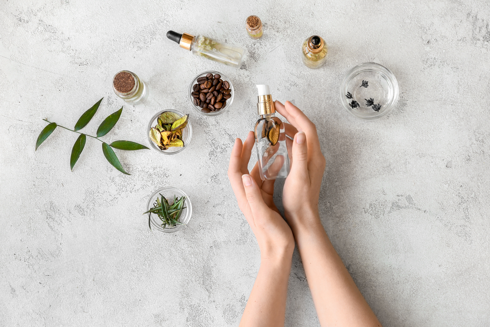 Mâini de femeie cu un recipient de parfum din sticlă alături de ingrediente naturale pe o suprafață de marmură albă cu gri.