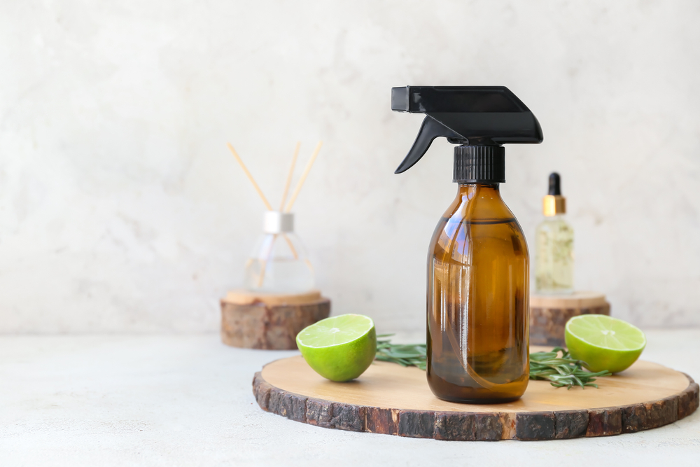 Parfum homemade: Un recipient din plastic maro transparent cu pulveriztor negru pe un fund rotund din lemn, alături de fructe de lime tăiate pe un fundal alb.