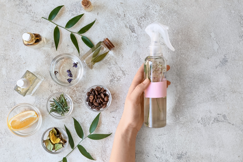 Parfum homemade: Mână de femmei ține un recipient transparent din plastic cu pulverizator cu un lichi sugestiv pentru odorizant de casă, alături de ingrediente naturale pentru parfumuri.