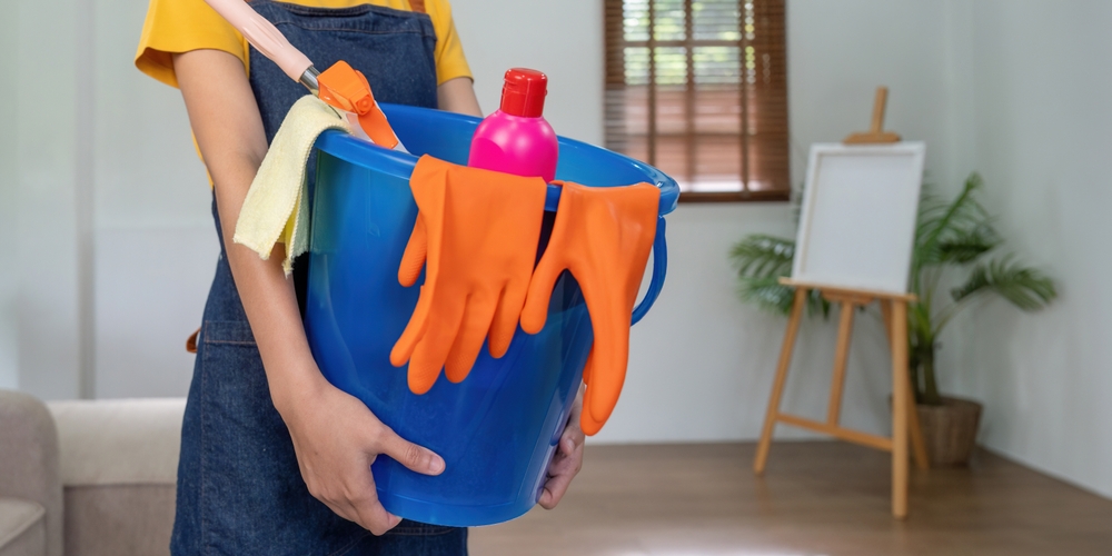 O femeie duce în brațe o găleată albastră cu accesorii pentru curățenie generală.