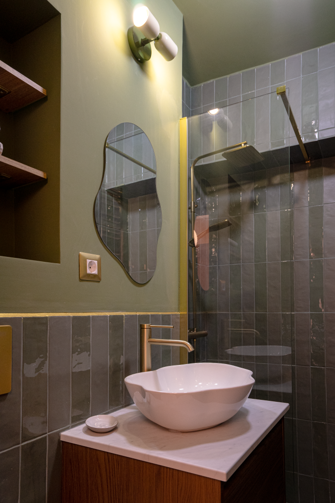 baie amenajată cu pereți și ceramică verde pădure cu oglindă cu formă neregulată, robinet auriu, lavoar alb cu formă neregulată și mască de chiuvetă din lemn