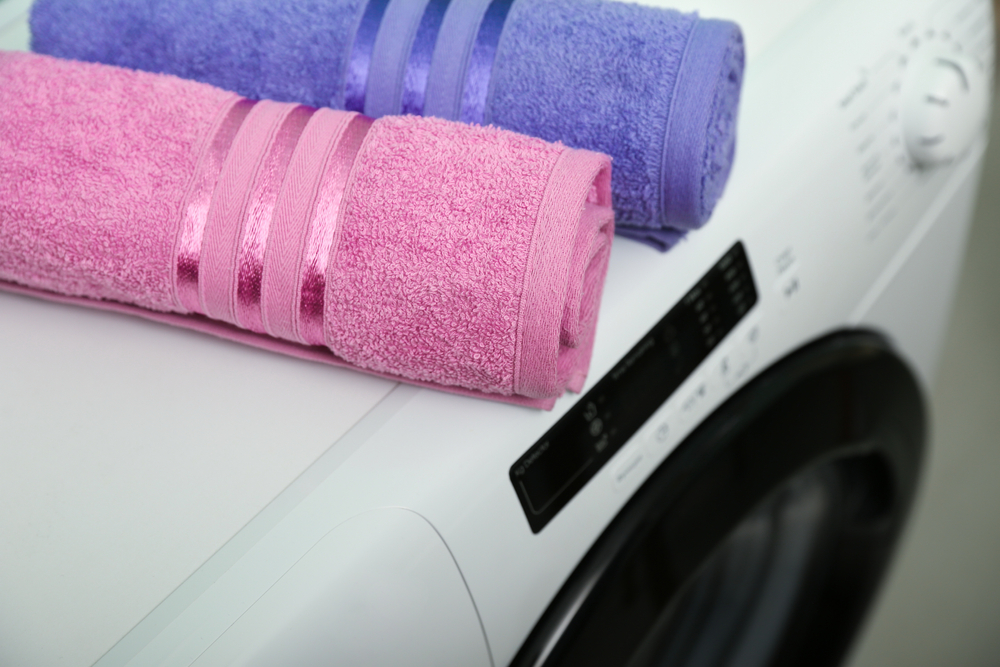 Un prosop de față roz și unul violet împăturite cilindric pe o mașină de spălat haine.