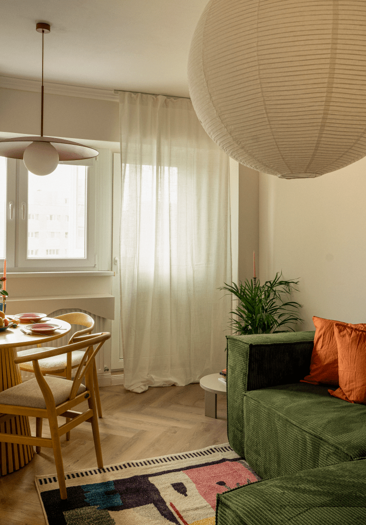 amenajare living cu pereti albi canapea verde perne decorative portocalii covor alb cu roz si nleumarin si masa cu scaune din lemn natur si tapiterie din boucle alb