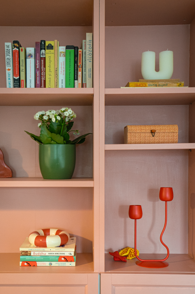 rafturi de biblioteca vopsite in roz decorate cu carti, lumanari, un ghiveci cu plante verzi si un sfesnic dublu rosu