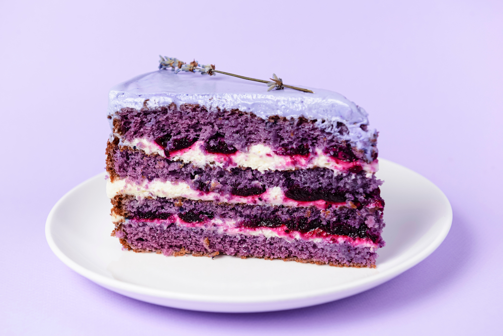 O felie de tort de lavandă cu mai multe straturi de cremă albă și mov, cu glazură lila și o floare de lavandă.