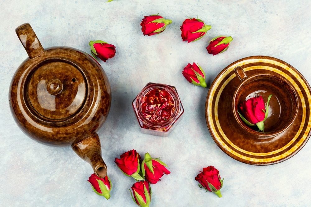 Flori comestibile naturale: Dulceașă de trandafiri alături de un ceainic și o ceașcă, pe o masă albă cu boboci și petale de trandafiri.