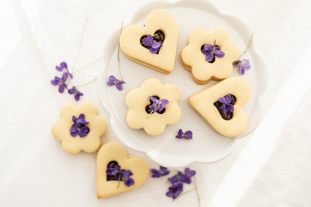 Biscuiți dulci în formă de inimă și flori umpluți cu gem și decorați cu violete pe o farfurie albă.