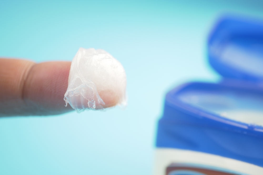Un deget cu vaselină cosmetică pe un fundal bleu, în depărtare cu recipientul de vaselină alb cu capac albastru.