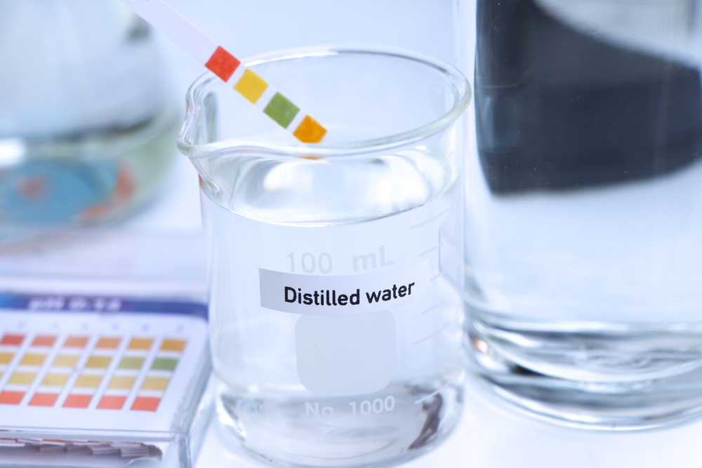 Un recipient transparent cu apă distlilată alături de un test din hârtie de pH.