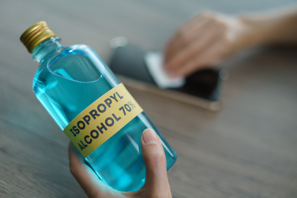 O persoană ține într-o mână o sticlă cu alcool izopropilic și cu cealaltă șterge cu o dischetă albă cu alcool un telefon mobil pe o suprafață din lemn.