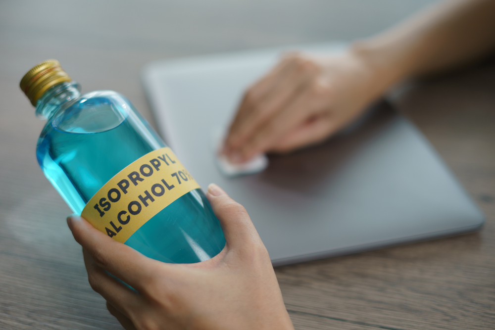 Alcool izopropilic la ce se folosește: O persoană ține într-o mână o sticlă de alcool izopropilic și cu cealaltă șterge un laptop închis