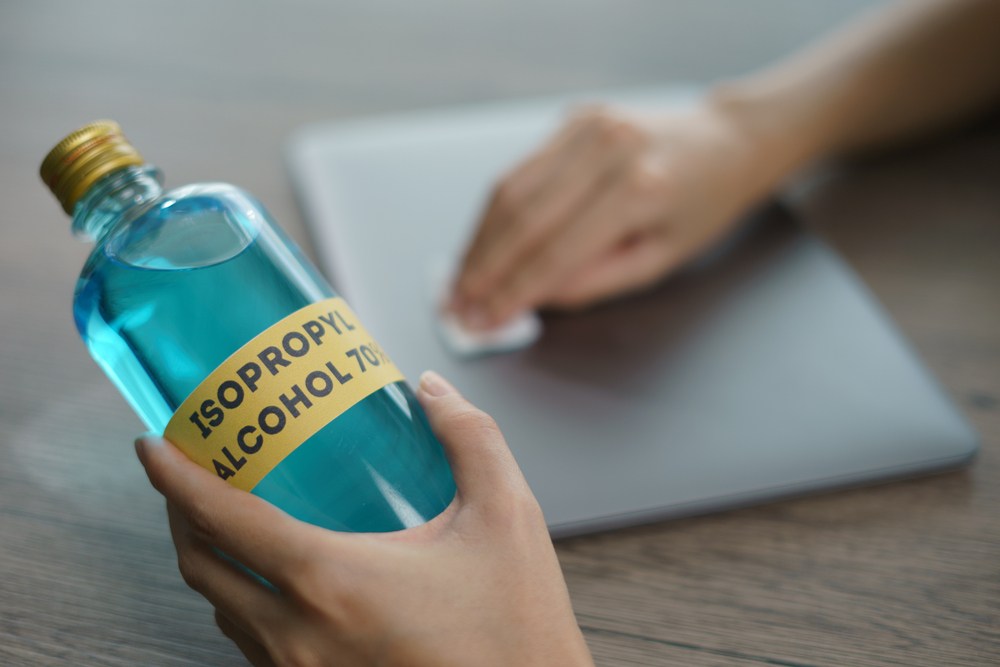 Alcool izopropilic la ce se folosește: O persoană ține într-o mână o sticlă de alcool izopropilic și cu cealaltă șterge un laptop închis